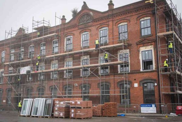 A LISTED BUILDING RENOVATION IN THE UK. The image depicts a Victorian brick warehouse building undergoing a sustainable retrofit, featuring scaffolding and the installation of new windows. This transformation highlights sustainable architecture examples, focusing on energy efficiency and the use of sustainable materials to minimize environmental impact.